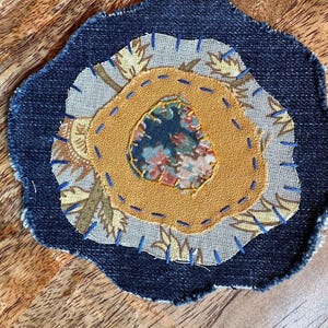 May include: A circular fabric patch featuring a dark blue denim base with frayed edges. The center displays layers of beige patterned fabric, a solid mustard yellow fabric, and a small floral print fabric, all secured with visible blue hand-stitched embroidery.