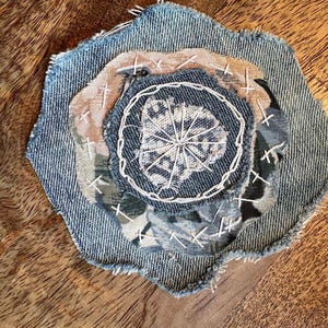 May include: A layered fabric applique featuring a blue denim base with frayed edges, topped with a circular piece of light brown fabric and a smaller blue denim center, all secured with white hand-stitched embroidery in a radial pattern.