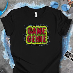 Könnte beinhalten: Schwarzes T-Shirt mit dem Schriftzug "GAME GENIE" in roter und grüner Pixeloptik, umrandet von gelben und blauen Blitzen. Das T-Shirt ist aus weichem Material.