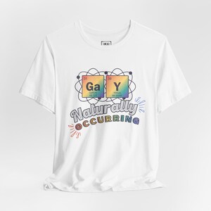 Op de afbeelding: Een wit T-shirt met korte mouwen en een afbeelding van twee elementen uit het periodiek systeem, Gallium en Yttrium, die het woord "GaY" vormen omringd door een atoom-symbool, met de tekst "Naturally OCCURRING" in een meerkleurig lettertype.
