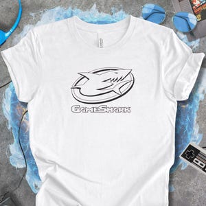 May include: White t-shirt featuring a black and white GameShark logo with a shark design inside an oval. The text "GameShark" is below the logo. The shirt is made of a soft material.