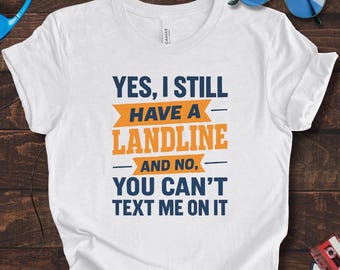 Peut inclure: T-shirt blanc avec le texte "YES, I STILL HAVE A LANDLINE - AND NO, YOU CAN'T TEXT ME ON IT" en bleu et orange. Le t-shirt a un col rond et des manches courtes. Des lunettes de soleil et un casque sont visibles en arrière-plan.
