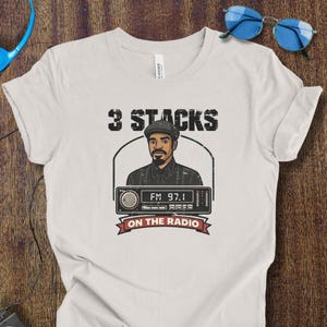 Op de afbeelding: Wit T-shirt met een afbeelding van een man en een radio. De tekst "3 STACKS" staat boven de afbeelding en "ON THE RADIO" staat eronder. De radio geeft "FM 97.1" weer.