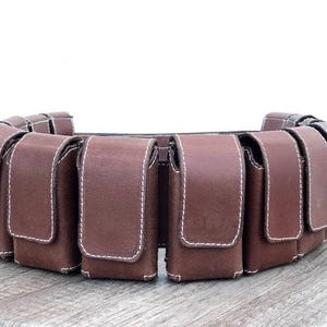 May include: A brown leather belt with multiple pouches. Each pouch has a flap closure and is stitched with white thread. The pouches are evenly spaced around the belt, designed to be worn around the waist. The belt is made of leather.