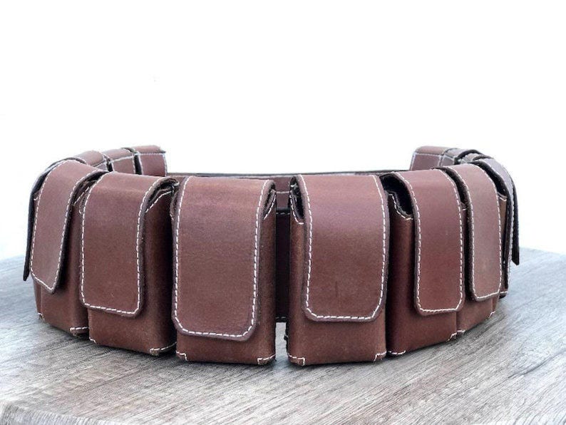 May include: A brown leather belt with multiple pouches. Each pouch has a flap closure and is stitched with white thread. The pouches are evenly spaced around the belt, designed to be worn around the waist. The belt is made of leather.