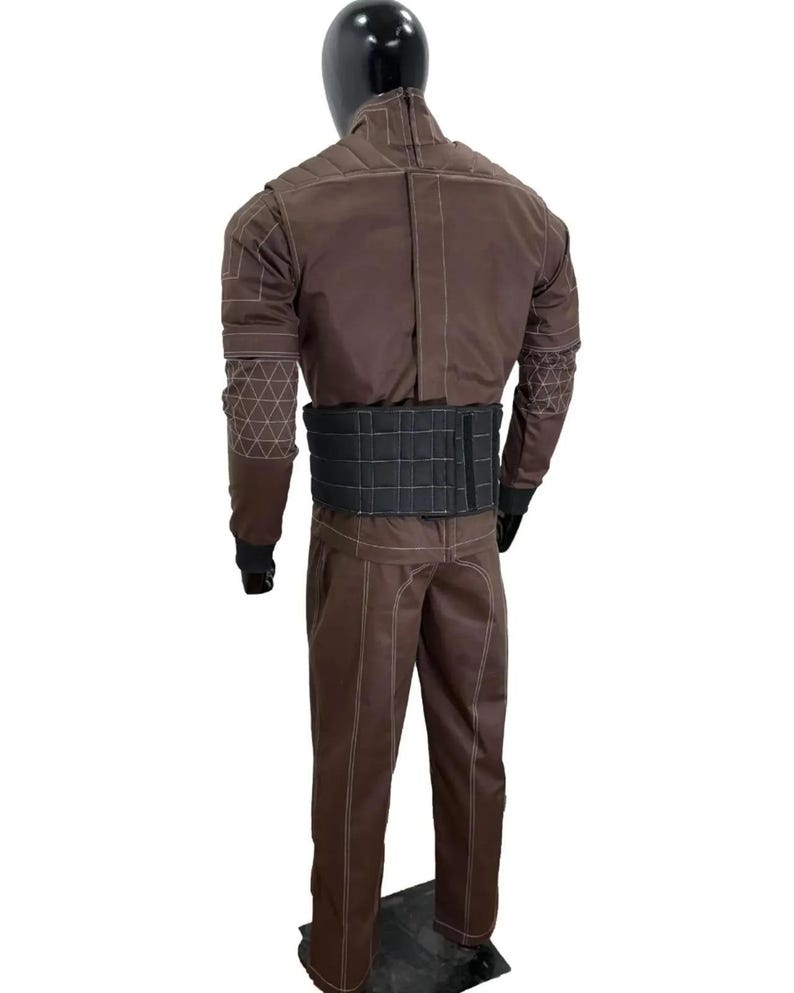 May include: Brown jumpsuit with white stitching and a black belt. The jumpsuit features geometric patterns on the sleeves and a high collar. The mannequin is standing on a black base.