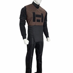 Puede incluir: Un atuendo completo en un maniqu&iacute;. El atuendo incluye un chaleco marr&oacute;n con un logotipo negro, una camisa negra de manga larga, pantalones negros con costuras blancas y un cuello alto negro. El atuendo tiene un dise&ntilde;o futurista.