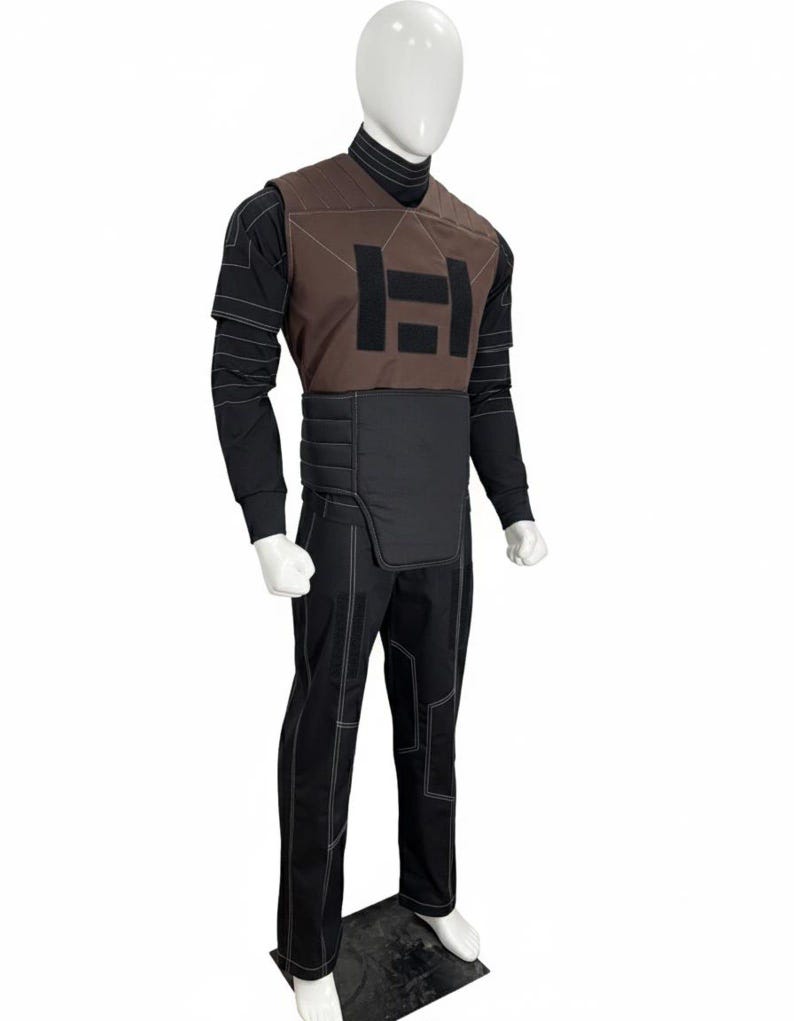 Puede incluir: Un atuendo completo en un maniqu&iacute;. El atuendo incluye un chaleco marr&oacute;n con un logotipo negro, una camisa negra de manga larga, pantalones negros con costuras blancas y un cuello alto negro. El atuendo tiene un dise&ntilde;o futurista.