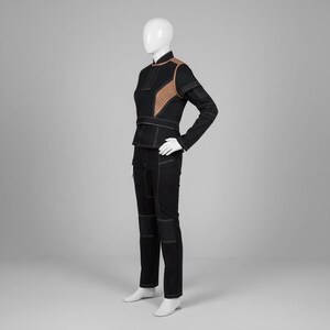 Puede incluir: Un conjunto negro de dos piezas sobre un maniqu&iacute;. La chaqueta tiene detalles en los hombros de color beige y costuras blancas. Los pantalones son negros con costuras blancas en las rodillas y los bolsillos. El conjunto se exhibe en un maniqu&iacute; blanco.