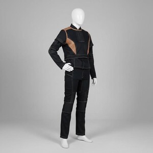 Puede incluir: Mono negro con detalles en los hombros color marr&oacute;n y costuras blancas. El conjunto incluye una chaqueta de manga larga y pantalones a juego. El maniqu&iacute; lleva zapatos blancos. El conjunto se muestra sobre un fondo gris.