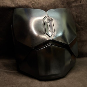 Puede incluir: Una pieza de armadura pectoral gris met&aacute;lico con un dise&ntilde;o geom&eacute;trico. La armadura tiene una costura vertical central y un detalle rectangular en la parte superior. La superficie refleja la luz, resaltando su forma y textura. El dise&ntilde;o sugiere un tema futurista o de ciencia ficci&oacute;n.