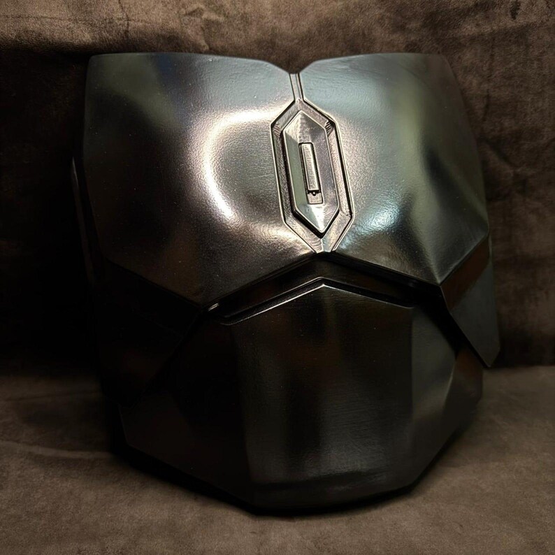 Puede incluir: Una pieza de armadura pectoral gris met&aacute;lico con un dise&ntilde;o geom&eacute;trico. La armadura tiene una costura vertical central y un detalle rectangular en la parte superior. La superficie refleja la luz, resaltando su forma y textura. El dise&ntilde;o sugiere un tema futurista o de ciencia ficci&oacute;n.