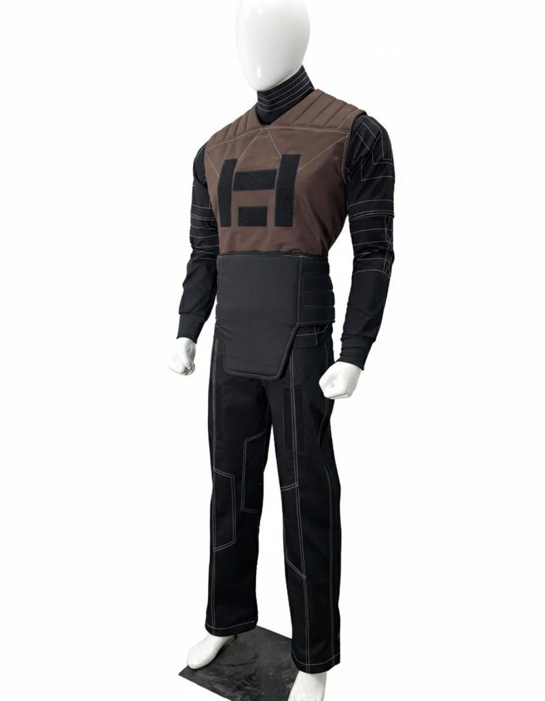 Puede incluir: Un atuendo completo que consta de un chaleco marr&oacute;n con un logotipo negro, una camisa negra de manga larga, pantalones negros con costuras blancas y un cintur&oacute;n negro. El atuendo se exhibe en un maniqu&iacute;.