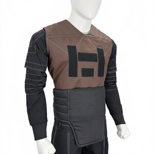 Puede incluir: Un chaleco t&aacute;ctico marr&oacute;n con un dise&ntilde;o "H" negro en el pecho, usado sobre una camisa de manga larga y pantalones negros. El chaleco tiene un dise&ntilde;o acolchado y se muestra en un maniqu&iacute;.