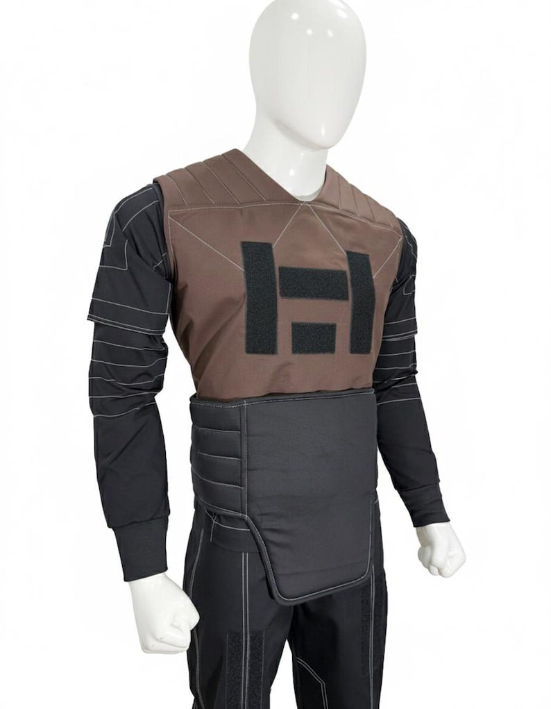 Puede incluir: Un chaleco t&aacute;ctico marr&oacute;n con un dise&ntilde;o "H" negro en el pecho, usado sobre una camisa de manga larga y pantalones negros. El chaleco tiene un dise&ntilde;o acolchado y se muestra en un maniqu&iacute;.