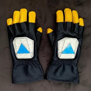 Puede incluir: Un par de guantes de cuero negro con las puntas de los dedos amarillas. Cada guante presenta un parche plateado con un diseño de triángulo azul. Guantes para conducir o correr.