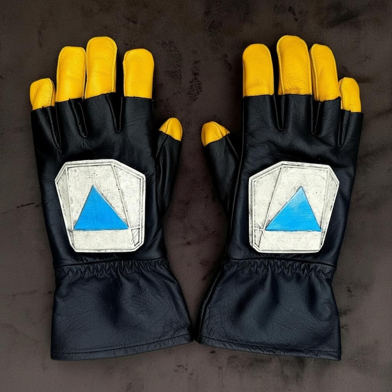 May include: A pair of black leather gloves with yellow fingertips. Each glove features a silver patch with a blue triangle design. The gloves are likely for driving or racing.