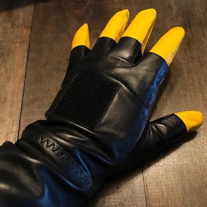 May include: Black leather glove with yellow fingertips. The glove features a black Velcro patch on the back of the hand and a stitched wrist. The fingers are a bright yellow color, contrasting with the black leather.