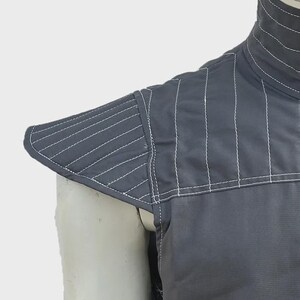 May include: Close-up of a gray garment with white stitching. The garment features a high collar and a padded shoulder piece with diagonal stitching. The fabric appears to be a durable material, suitable for protection or layering.