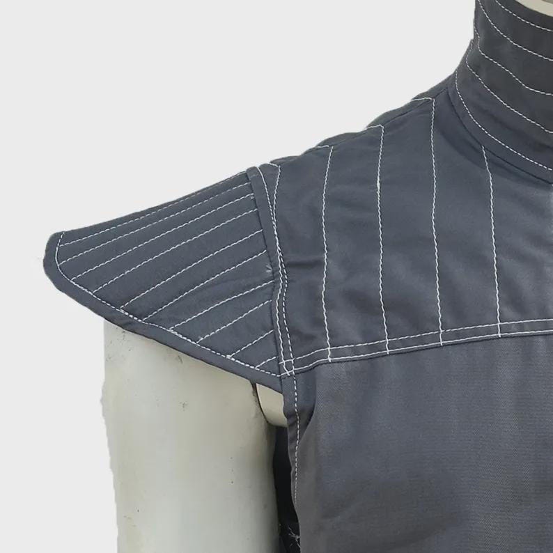 May include: Close-up of a gray garment with white stitching. The garment features a high collar and a padded shoulder piece with diagonal stitching. The fabric appears to be a durable material, suitable for protection or layering.