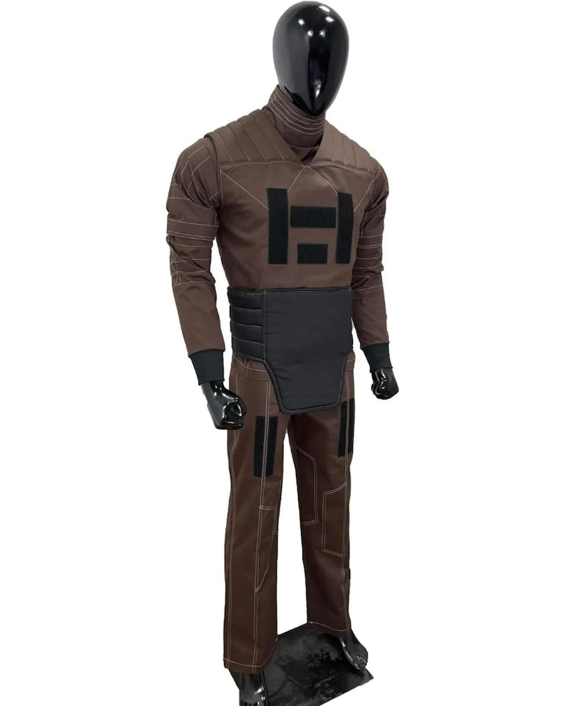 May include: Brown jumpsuit with black accents, displayed on a mannequin. The outfit features a high collar, long sleeves, and a black padded belt. The chest has a large black graphic. The pants have black patches and white stitching.