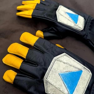 May include: A pair of black leather gloves with yellow fingertips and a silver emblem with a blue triangle. The gloves are lying on a brown surface.