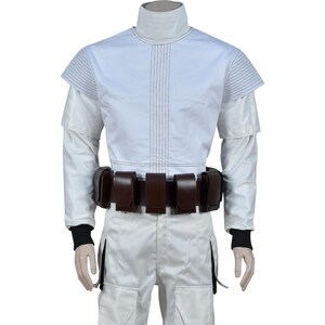 May include: White costume with a high collar and a brown belt with pouches. The top has a short-sleeved design with vertical stitching down the front. The pants are white with a pocket detail. Black cuffs are visible at the wrists.