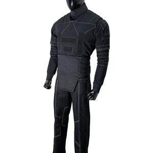 Puede incluir: Mono t&aacute;ctico negro con costuras blancas. El mono presenta un cuello alto, mangas largas y un dise&ntilde;o ajustado. La prenda se exhibe en un maniqu&iacute; negro, mostrando sus paneles estructurados y &aacute;reas reforzadas.