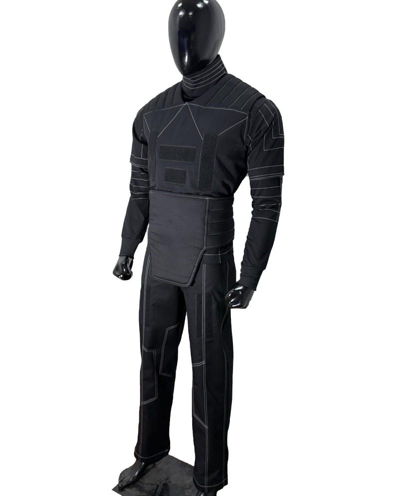 Puede incluir: Mono t&aacute;ctico negro con costuras blancas. El mono presenta un cuello alto, mangas largas y un dise&ntilde;o ajustado. La prenda se exhibe en un maniqu&iacute; negro, mostrando sus paneles estructurados y &aacute;reas reforzadas.