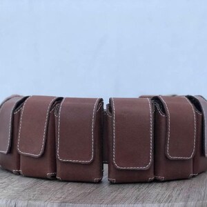 May include: A brown leather belt featuring several rectangular pouches with flap closures, stitched with white thread. The belt, designed for waist wear, is displayed on a wooden surface. The pouches are likely for carrying small items.