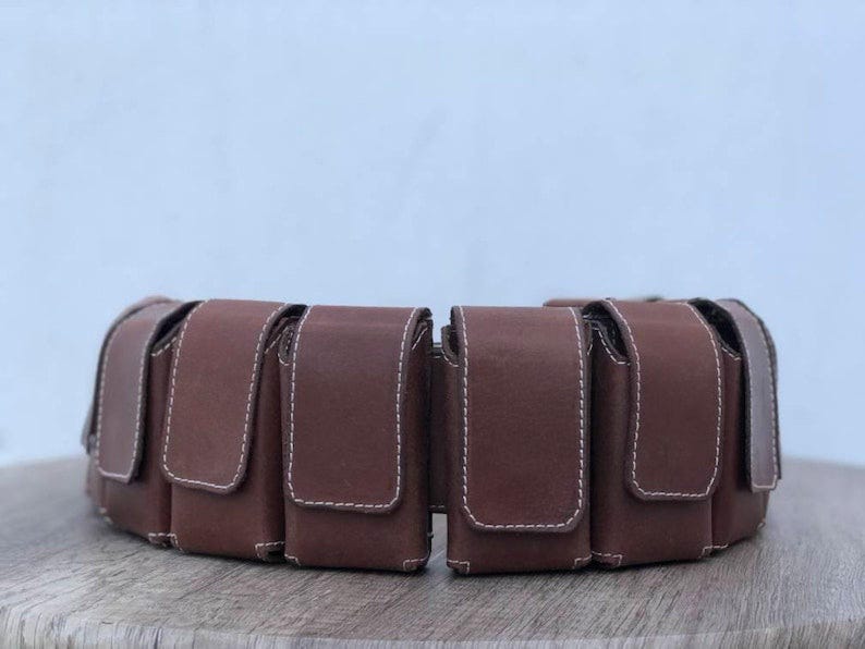 May include: A brown leather belt featuring several rectangular pouches with flap closures, stitched with white thread. The belt, designed for waist wear, is displayed on a wooden surface. The pouches are likely for carrying small items.