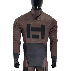 May include: Brown protective suit with black accents, displayed on a mannequin. The suit features a high collar, padded shoulders, and a black panel across the midsection. Black patches and cuffs complete the design. The mannequin has a black head.