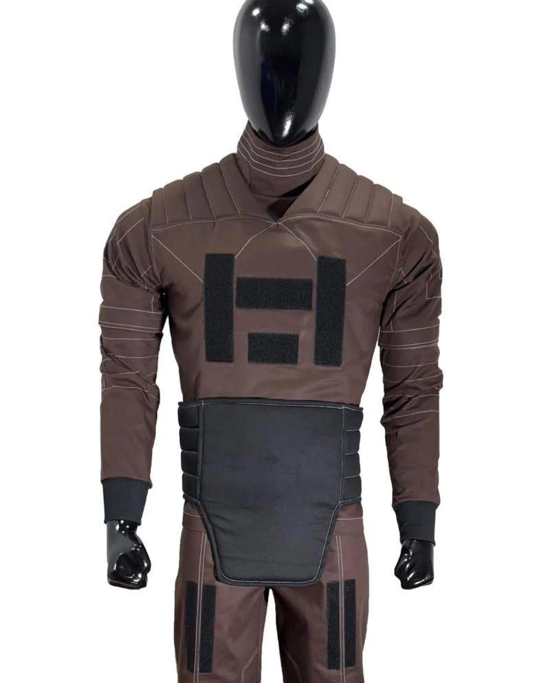 May include: Brown protective suit with black accents, displayed on a mannequin. The suit features a high collar, padded shoulders, and a black panel across the midsection. Black patches and cuffs complete the design. The mannequin has a black head.