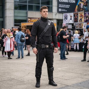 Pode incluir: Um traje t&aacute;tico preto composto por uma blusa de manga comprida com gola alta e costuras geom&eacute;tricas, cal&ccedil;as a condizer, um cinto utilit&aacute;rio com bolsas e coldre, uma al&ccedil;a diagonal com fivela met&aacute;lica e botas pretas.