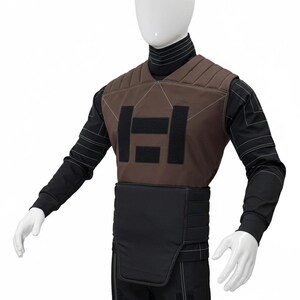 Puede incluir: Un chaleco t&aacute;ctico marr&oacute;n y negro con un dise&ntilde;o geom&eacute;trico y la letra "H" en la parte delantera. El chaleco se lleva sobre una camisa negra de manga larga y pantalones. El maniqu&iacute; lleva un cuello alto negro.