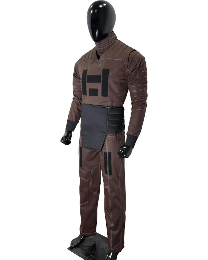May include: A brown costume with a high collar, long sleeves, and matching pants. The costume features a black panel across the midsection and the letter "H" on the chest. White stitching accents the seams. The costume is displayed on a black mannequin.