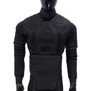 Puede incluir: Chaleco t&aacute;ctico negro con detalles de costuras blancas, exhibido en un maniqu&iacute;. El chaleco presenta m&uacute;ltiples paneles, incluyendo protecci&oacute;n para hombros y pecho, y mangas largas. El maniqu&iacute; lleva pantalones negros.