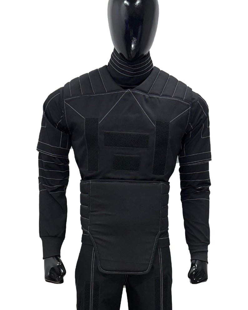 Puede incluir: Chaleco t&aacute;ctico negro con detalles de costuras blancas, exhibido en un maniqu&iacute;. El chaleco presenta m&uacute;ltiples paneles, incluyendo protecci&oacute;n para hombros y pecho, y mangas largas. El maniqu&iacute; lleva pantalones negros.
