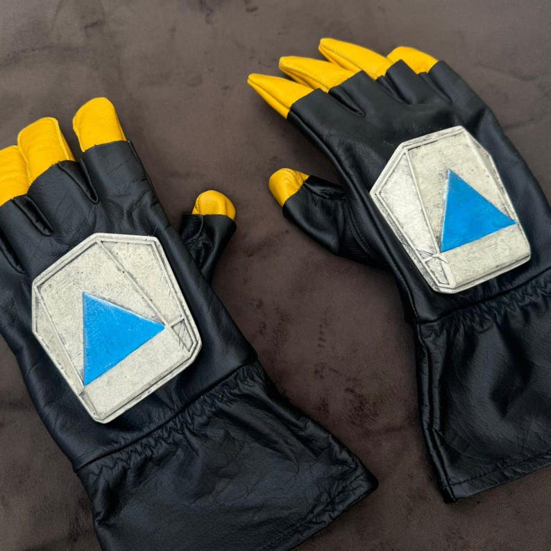 Puede incluir: Un par de guantes negros con las puntas de los dedos amarillas. Cada guante presenta un emblema plateado con un tri&aacute;ngulo azul. Los guantes parecen estar hechos de cuero o material similar.