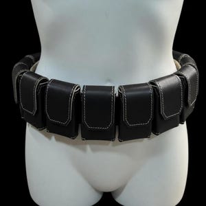 May include: A black leather utility belt with multiple pouches, each outlined with white stitching. The belt is displayed on a white mannequin, showcasing its design and construction. The pouches are evenly spaced around the belt's circumference.
