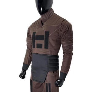 May include: Brown jumpsuit with black accents, displayed on a mannequin. The outfit features a high collar, quilted panels, and a black belt. The chest area has a large black square design. The sleeves have black cuffs.