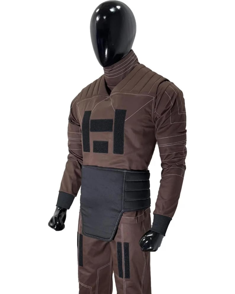 May include: Brown jumpsuit with black accents, displayed on a mannequin. The outfit features a high collar, quilted panels, and a black belt. The chest area has a large black square design. The sleeves have black cuffs.