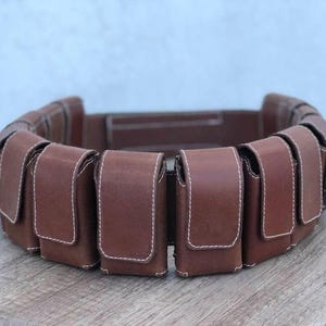 May include: A brown leather belt featuring several rectangular pouches. Each pouch is stitched with white thread and has a flap closure. The belt is designed to be worn around the waist.