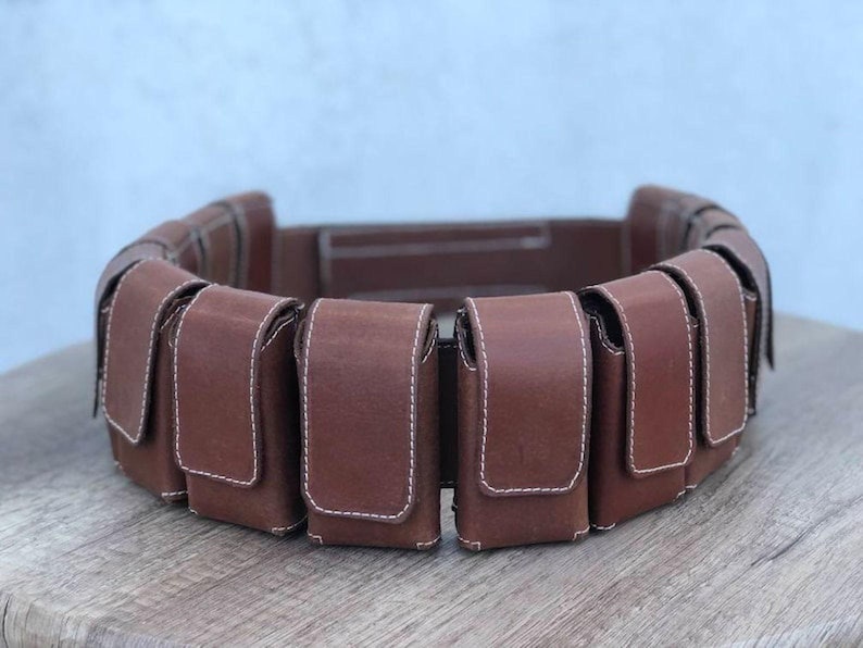 May include: A brown leather belt featuring several rectangular pouches. Each pouch is stitched with white thread and has a flap closure. The belt is designed to be worn around the waist.