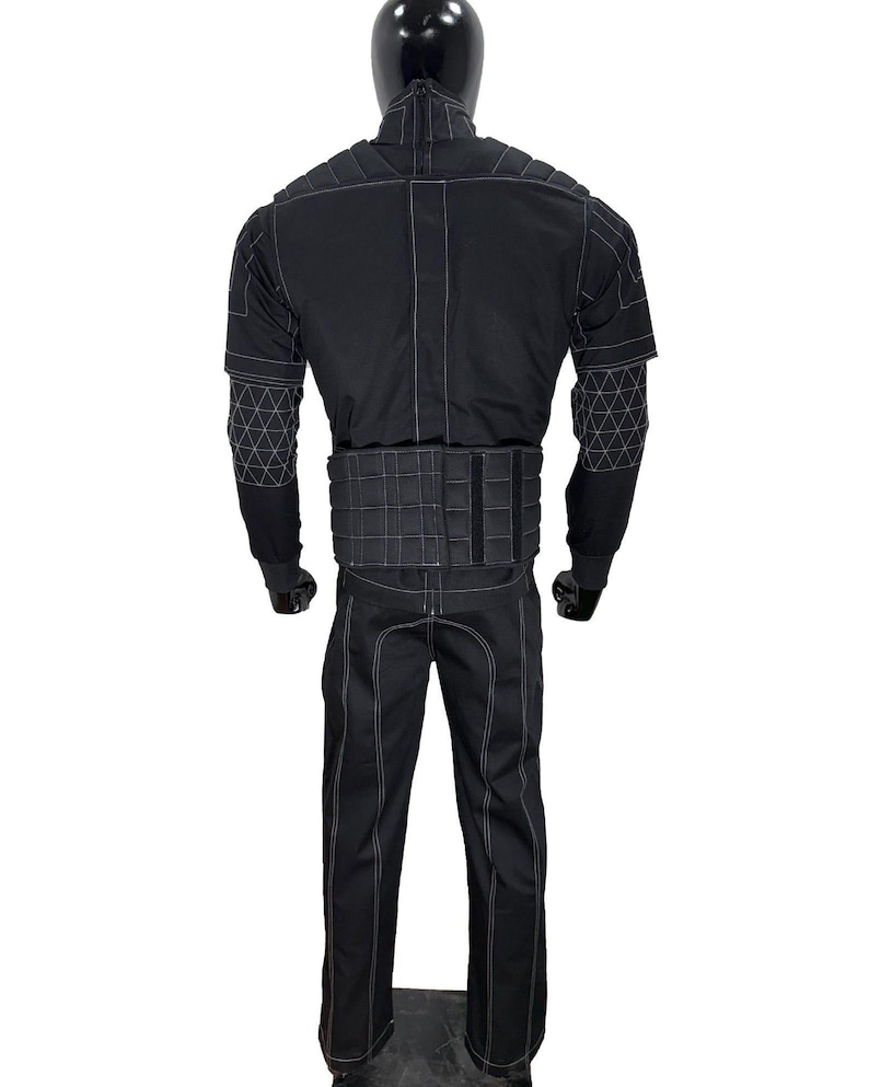 Puede incluir: Traje t&aacute;ctico negro exhibido en un maniqu&iacute;. El traje presenta costuras blancas que delinean las costuras y patrones geom&eacute;tricos en las mangas. El conjunto incluye una chaqueta, pantalones y un componente similar a un cintur&oacute;n en la cintura. El maniqu&iacute; tiene una cabeza negra.