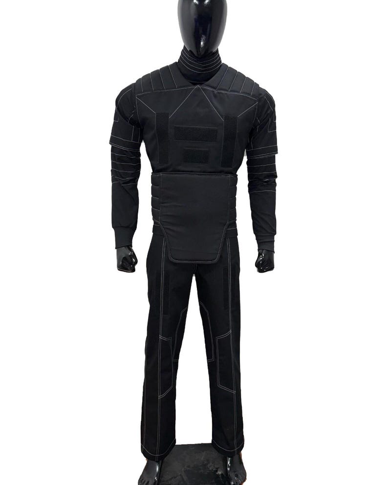 Puede incluir: Traje protector negro exhibido en un maniqu&iacute;. El traje incluye una parte superior de manga larga, pantalones y un cuello alto. Las costuras blancas delinean los paneles y las costuras del traje. El maniqu&iacute; tiene cabeza y manos negras.