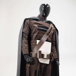 Puede incluir: Un disfraz marr&oacute;n y negro exhibido en un maniqu&iacute;. El disfraz incluye una parte superior de manga larga, pantalones, un cintur&oacute;n, una correa para el hombro y una capa gris. La parte superior tiene detalles en negro y patrones geom&eacute;tricos en las mangas.