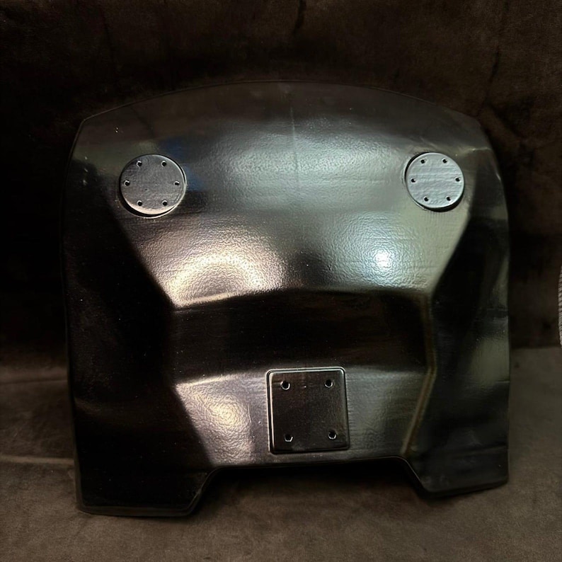 Puede incluir: Un componente de pl&aacute;stico negro moldeado con una superficie texturizada. Cuenta con dos fijaciones met&aacute;licas circulares y una placa rectangular con cuatro orificios para tornillos. El objeto tiene un dise&ntilde;o geom&eacute;trico con bordes angulados.