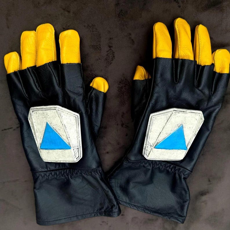 May include: A pair of black gloves with yellow fingertips. Each glove has a silver patch with a blue triangle. The gloves are made of leather and appear to be new.