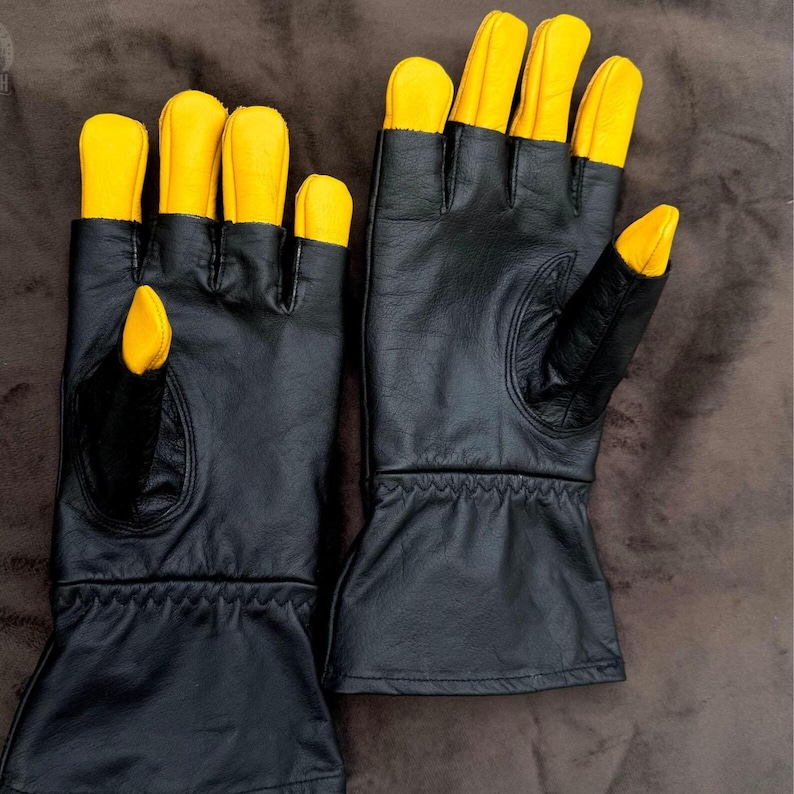 May include: A pair of black leather gloves with bright yellow fingertips. The gloves have a classic design with a wrist cuff and are likely for work or protection. The gloves are shown on a brown surface.